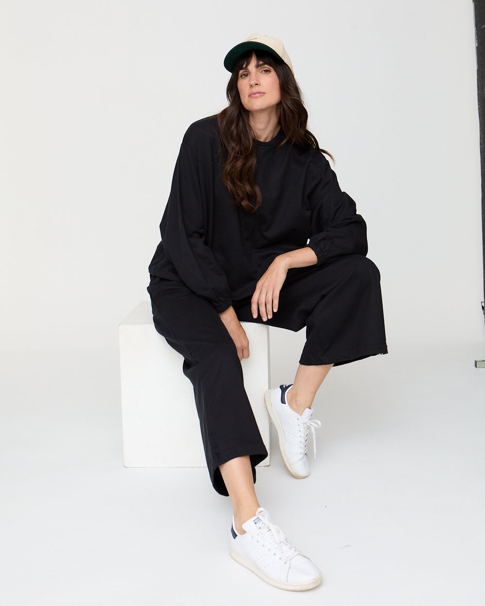 Woman wearing a black outfit with white sneakers sitting on a white chair against a white background
