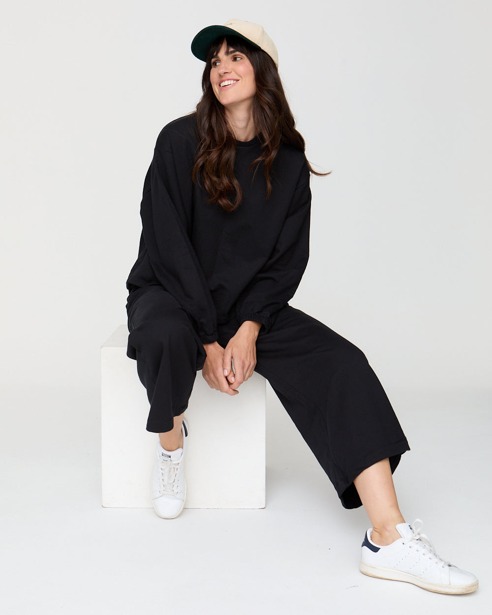 Woman wearing a black outfit with white sneakers sitting on a white stool against a white background