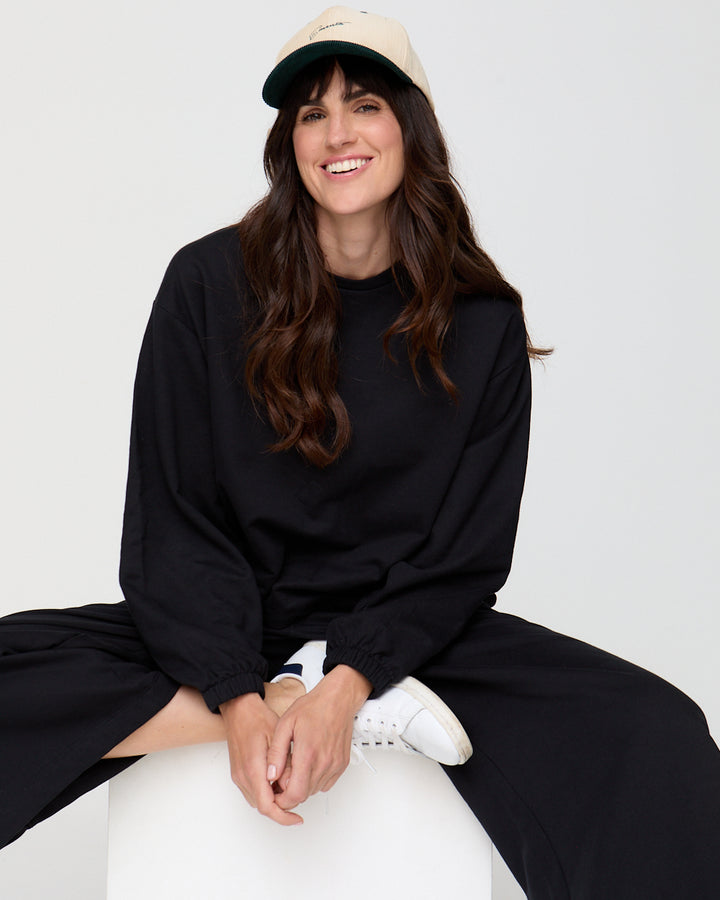 Woman wearing a black outfit and beige cap sitting on a white stool against a white background