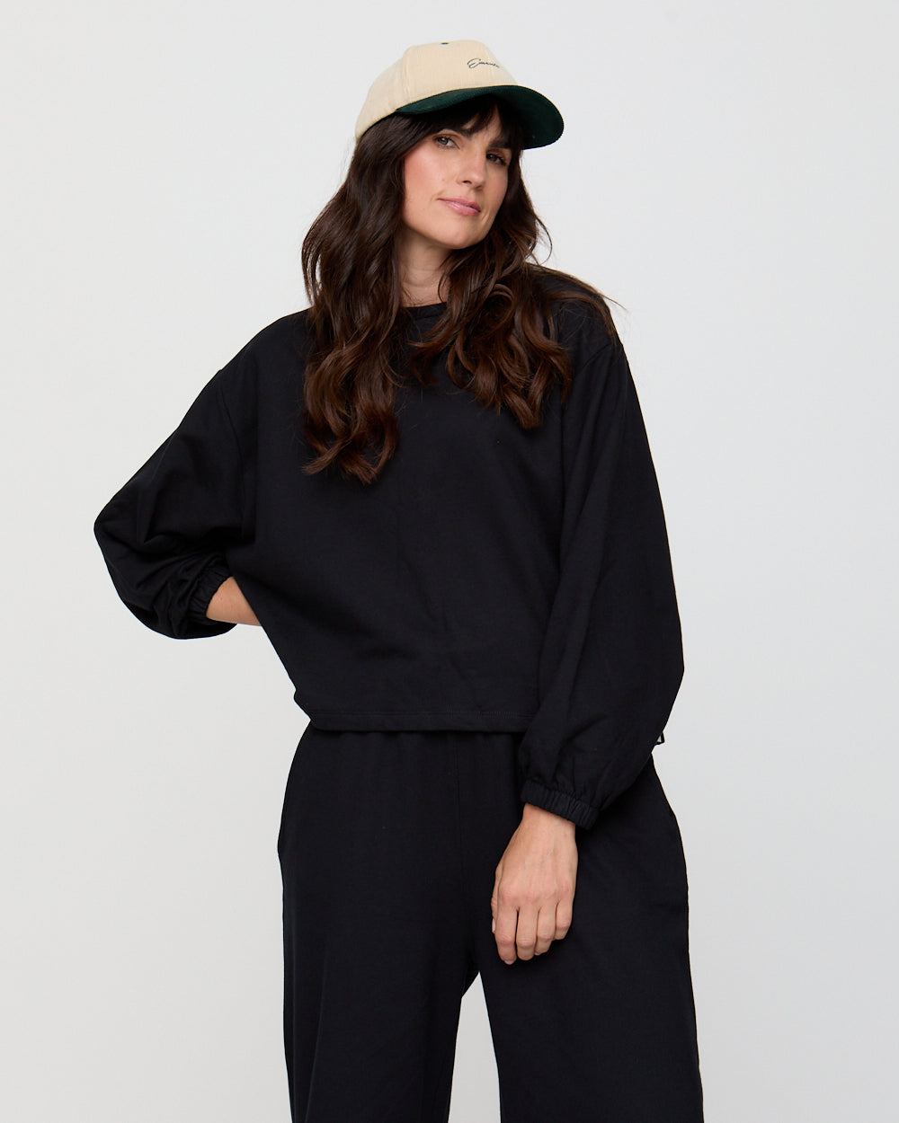 Woman wearing a black sweatshirt and pants with a beige cap on a light gray background