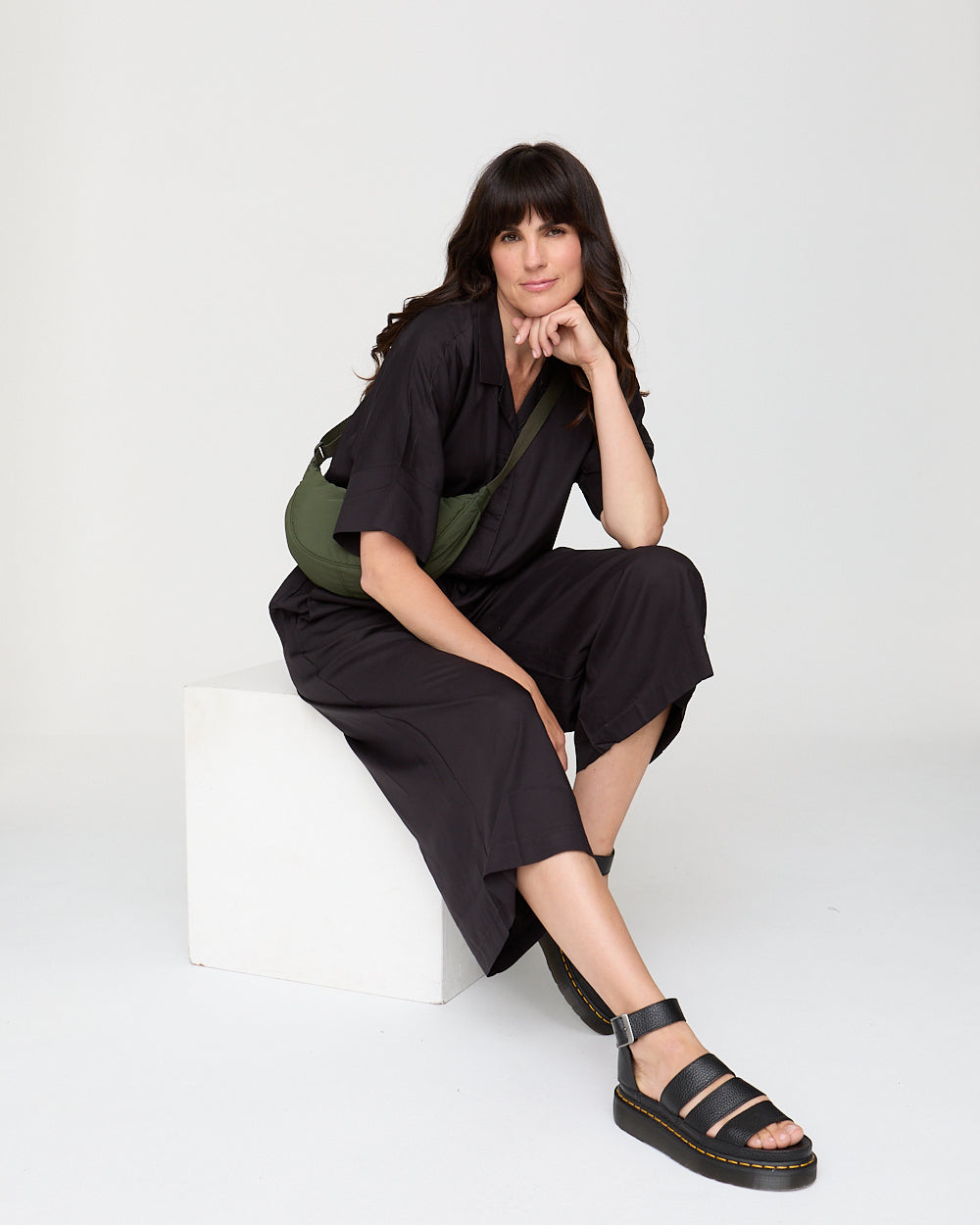 Woman in black outfit with green bag sitting on a white chair against a white background