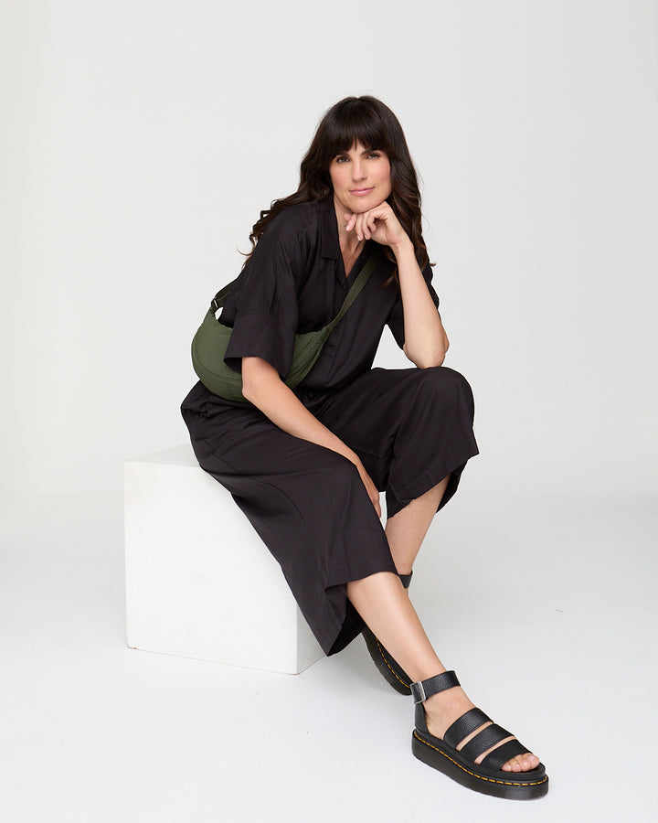 Woman in black outfit with green bag sitting on a white chair against a white background