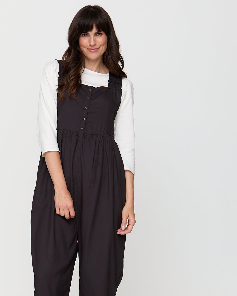 Woman wearing a black pinafore dress over a white shirt on a plain background