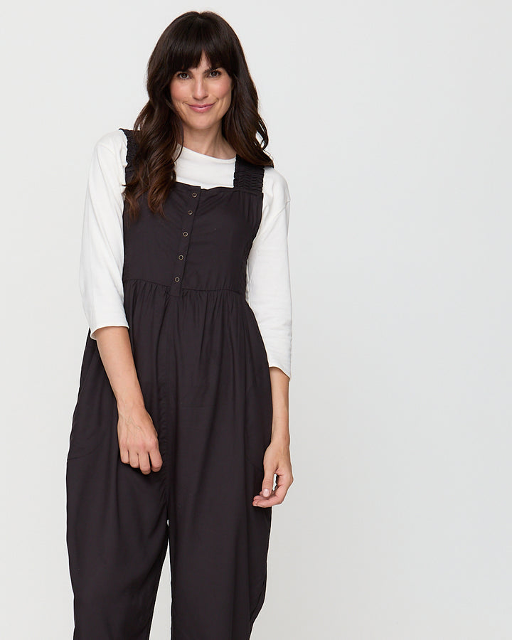Woman wearing a black pinafore dress over a white shirt on a plain background