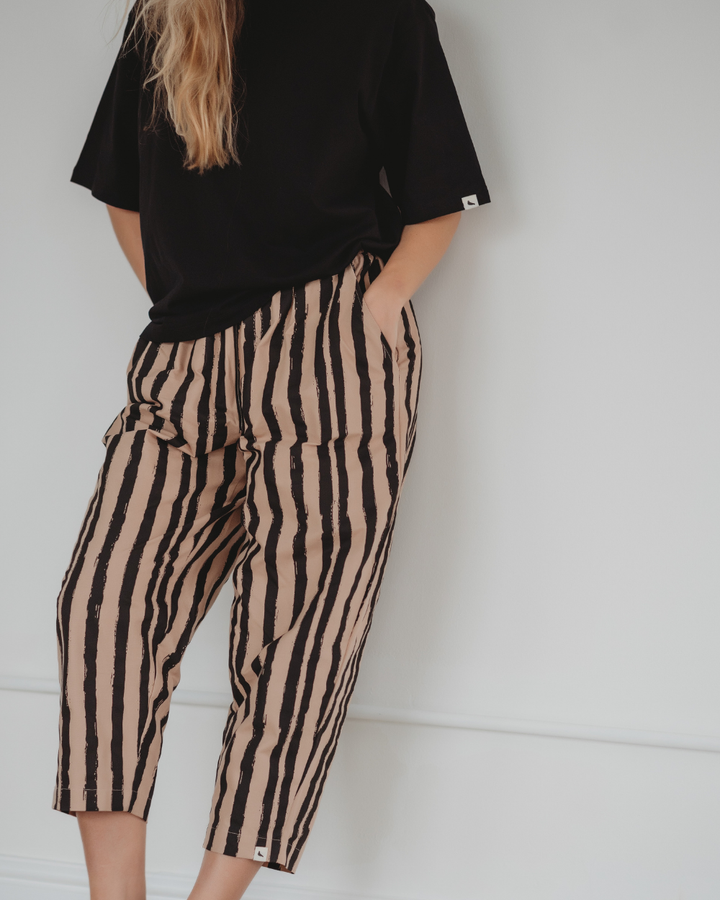 A person with long, wavy hair stands against a white background, wearing a black t-shirt and Kate Painted Stripe Trousers in beige with vertical black stripes and a barrel leg fit. Their hands rest in their pockets, reflecting a commitment to sustainable fashion through organic cotton.