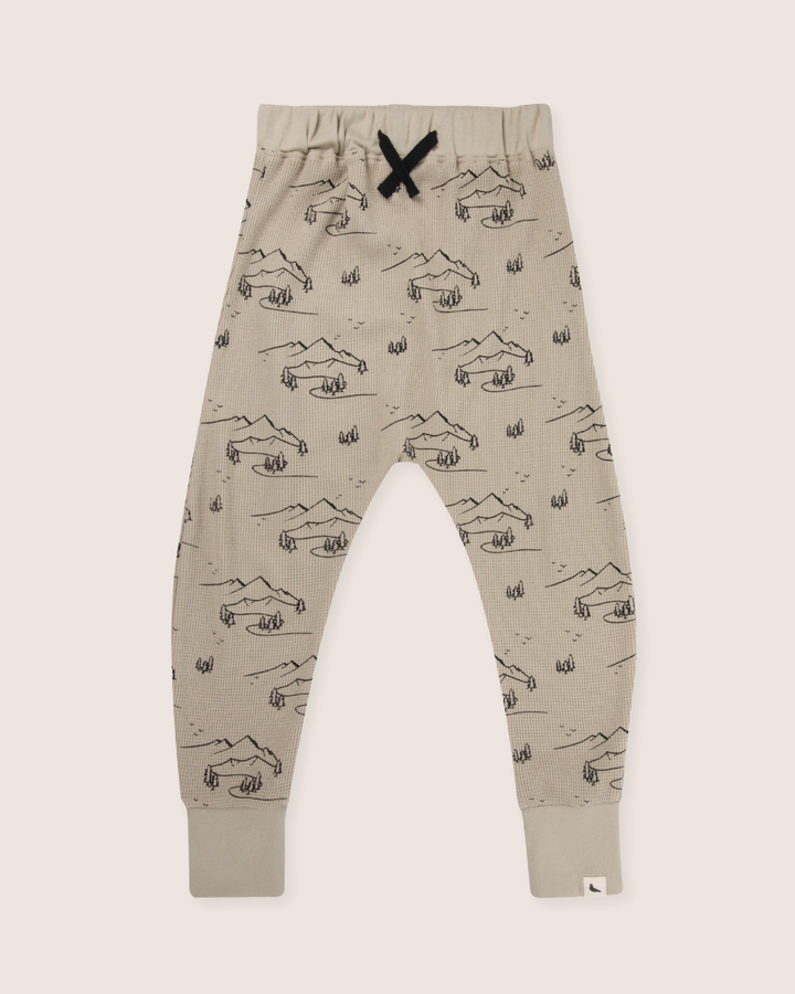 The Wilderness Joggers are beige, made from organic cotton with a charming print of black mountains and trees, featuring a stylish black drawstring at the waist.