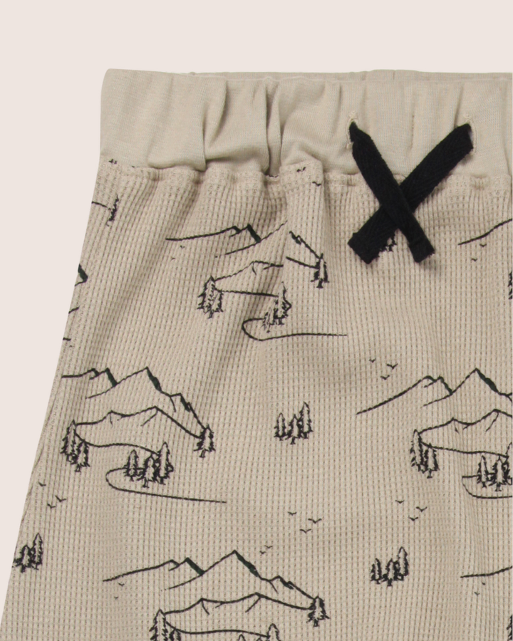 Close-up of Wilderness Joggers for kids in beige, showcasing a wilderness print with mountains and trees. These organic cotton pants feature an elastic waistband with a black drawstring bow and a textured, waffle-like fabric appearance.