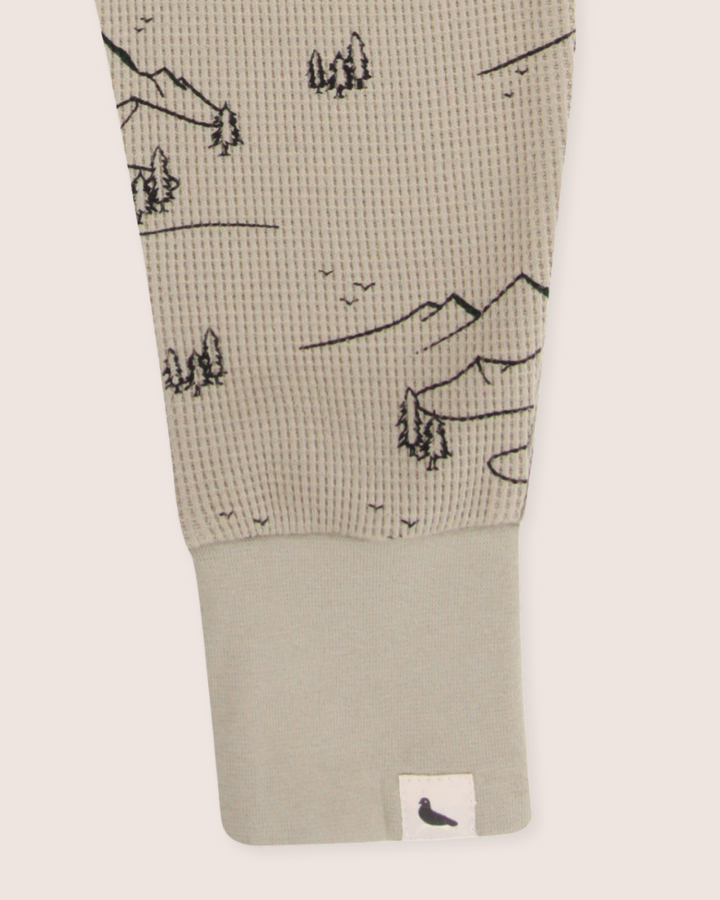 Close-up of the Wilderness Joggers sleeve with textured beige organic cotton fabric, displaying black mountain and tree prints. The smooth cuff has a small bird silhouette tag, ideal for kids seeking an adventure-ready look.