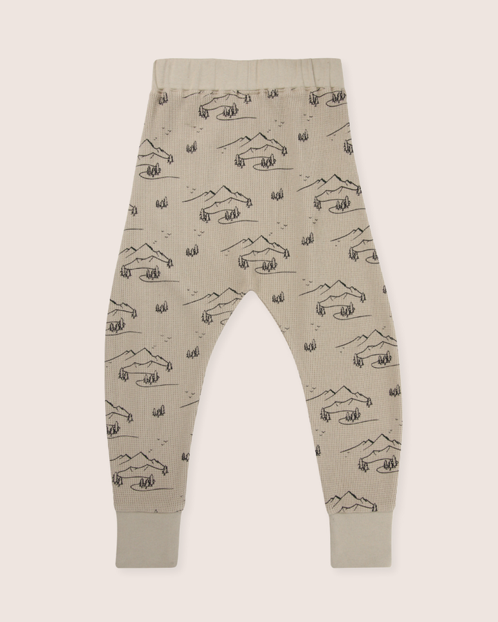 The Wilderness Joggers are beige toddler pants made from organic cotton, featuring an elastic waistband and cuffs, adorned with a playful doodle-style wilderness print of mountains, trees, and tents.