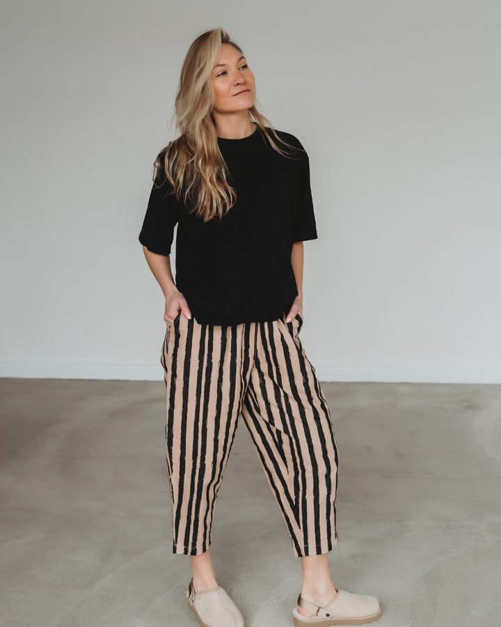 In a minimalist room, a person with long hair wears a black shirt and Kate Painted Stripe Trousers, featuring beige fabric with vertical black stripes. Crafted from organic cotton for sustainability, they pair the outfit with tan shoes, hands in pockets and gaze to the side with relaxed ease.