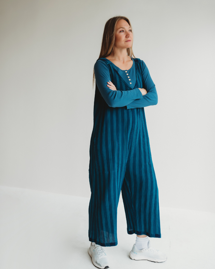 A confident woman stands in a minimal white room wearing the Grace Check & Stripe Reversible Navy Jumpsuit made from organic cotton and white sneakers, her arms folded as she gazes upwards, reflecting a sustainable wardrobe choice.