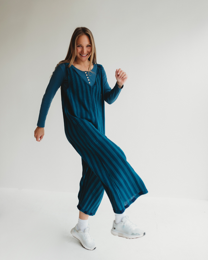 A woman joyfully kicks one leg up in a Grace Check & Stripe Reversible Navy Jumpsuit, her smile wide and infectious. Clad in this organic cotton outfit with white sneakers, her long hair flows freely as she strikes a playful pose against a plain white backdrop, embodying fun and movement.