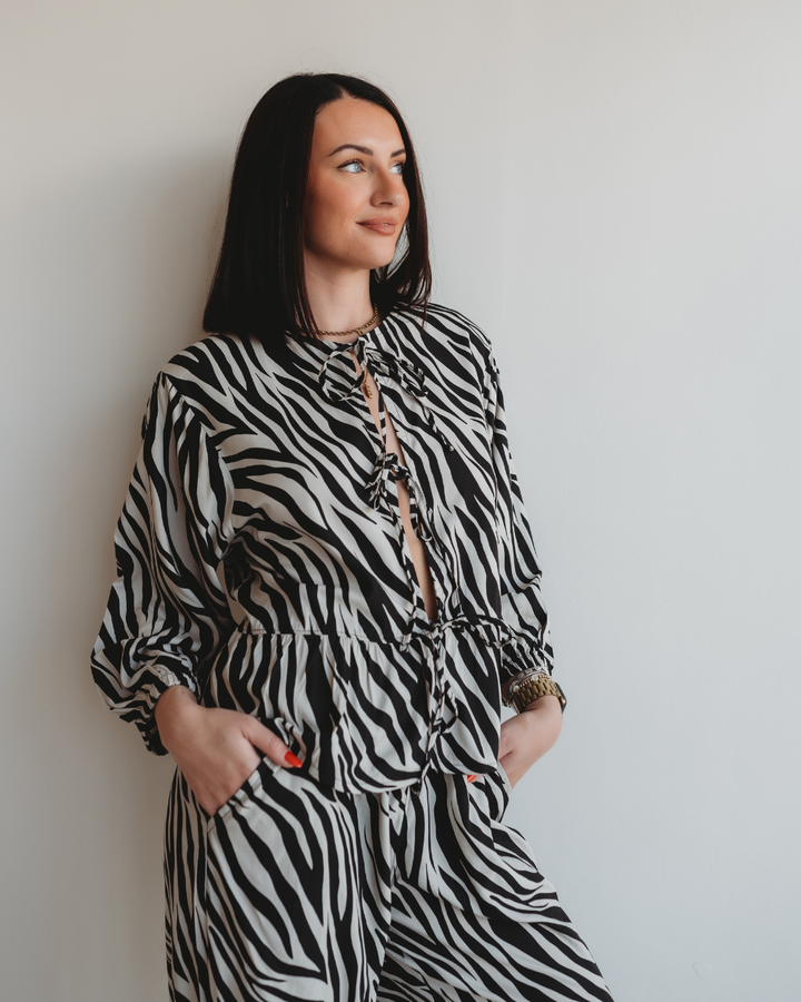 A woman with long dark hair leans against a white wall, hands in the pockets of her zebra-patterned Blake Zebra Print Tie Blouse Top. She gazes to the side with a slight smile.