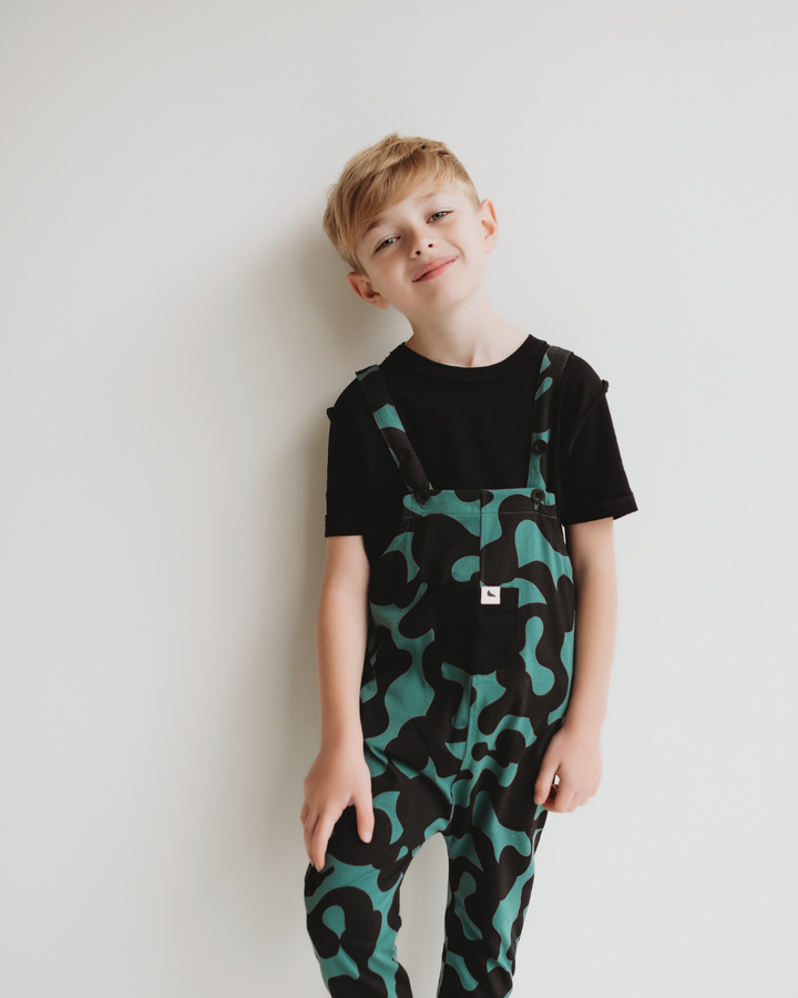 A young child with light hair, wearing Misty Print Dungarees made from organic cotton jersey and a black t-shirt, stands smiling against a white wall in a relaxed posture.