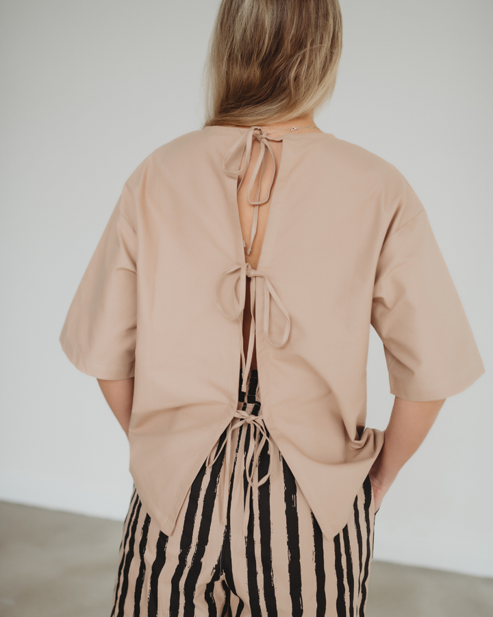 A person with long blond hair wears a Doris Tie Back Clay Top in beige, featuring an open back tied with knots, paired with striped black and beige pants. Made from organic cotton, the outfit pops in a minimalistic indoor setting as they face away from the camera.