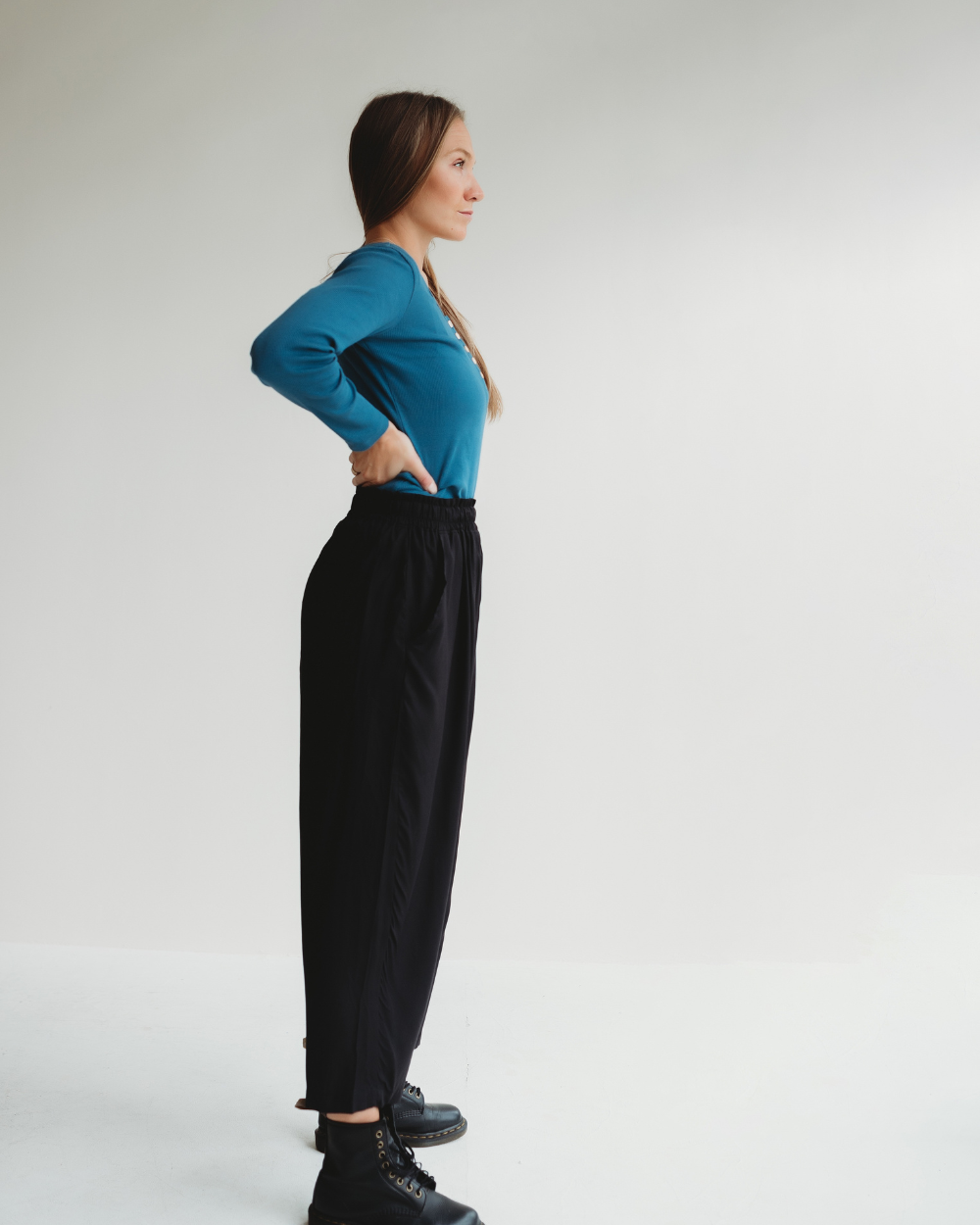 A woman stands in profile against a plain background, showcasing her sustainable wardrobe with a teal long-sleeve top made from organic cotton. Her look is completed with Betty Black Viscose Trousers and boots, as she confidently poses with hands on hips and elegantly braided hair.