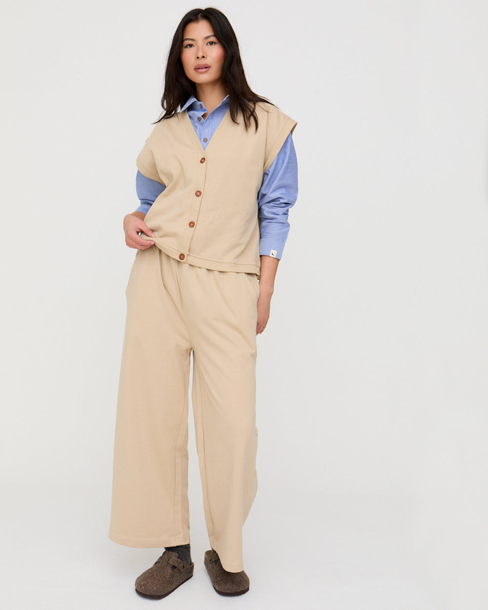 Woman wearing a beige vest over a blue shirt with beige pants on a white background