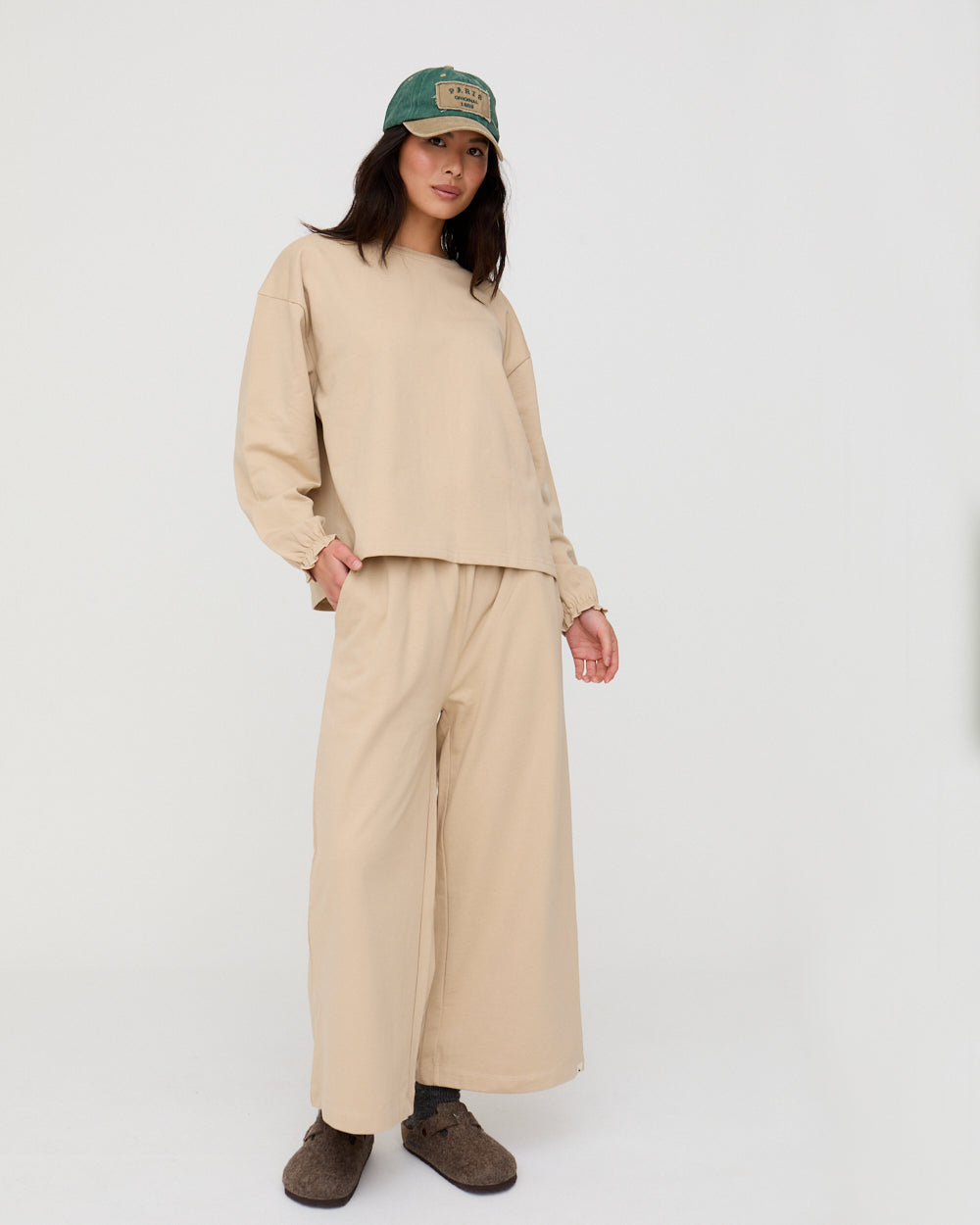 Person wearing a beige outfit with wide-leg pants and a cap on a white background