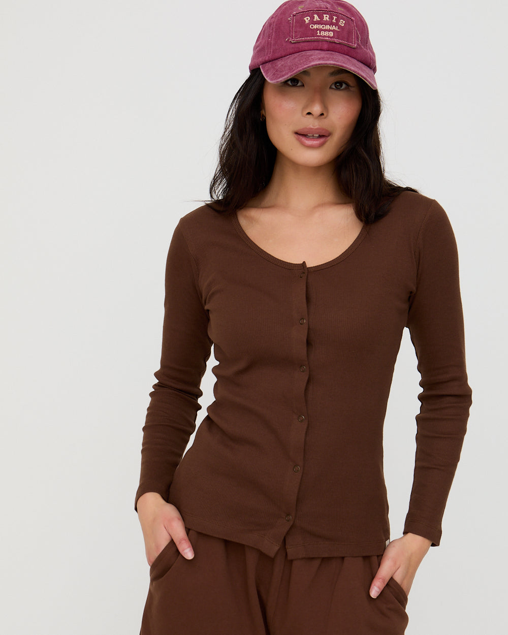 Woman wearing a brown long-sleeve top and pants with a purple cap on a light gray background