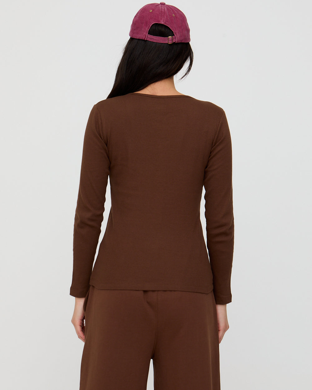 Person wearing a brown long-sleeve top and pants with a maroon cap on a white background