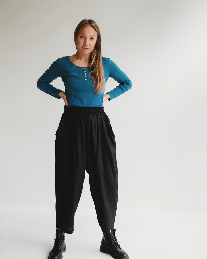 A person with long hair stands against a white backdrop, showcasing a sustainable wardrobe. They wear a blue long-sleeve top with buttons and Betty Black Viscose Trousers, both in contemporary drape style. Black boots complete the look as they pose relaxed, hands on hips.