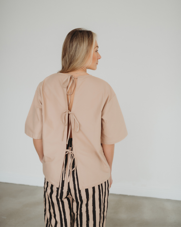 A person with long hair, standing indoors and facing away, wears the Doris Tie Back Clay Top made from organic cotton, characterized by a back opening tied with strings. This beige woven top is paired with striped black and white pants against a plain, light-colored background.