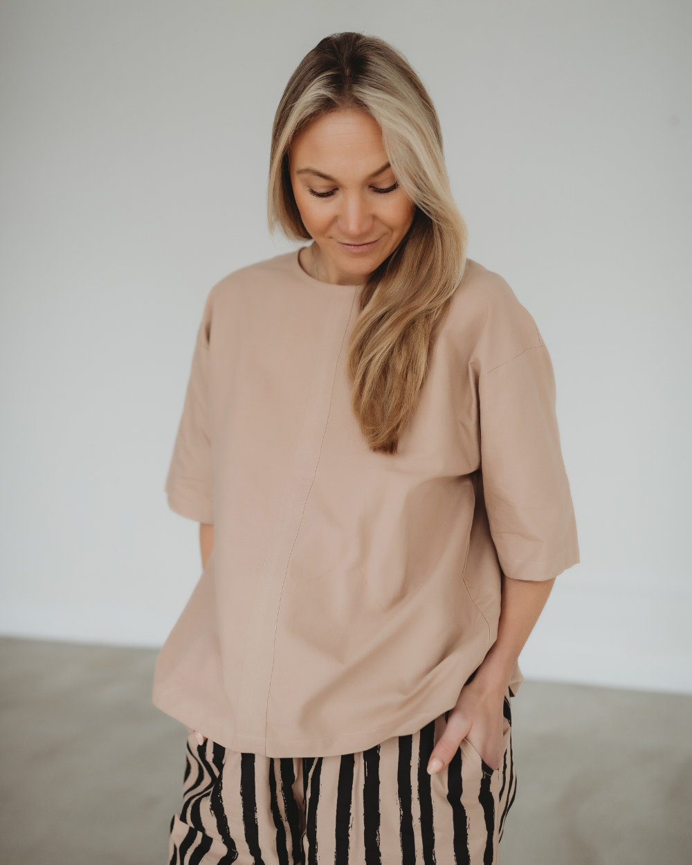 A woman with long blonde hair wears a versatile beige Doris Tie Back Clay Top and striped pants in a light-colored room. She looks down with a subtle smile, one hand casually tucked in her pocket.