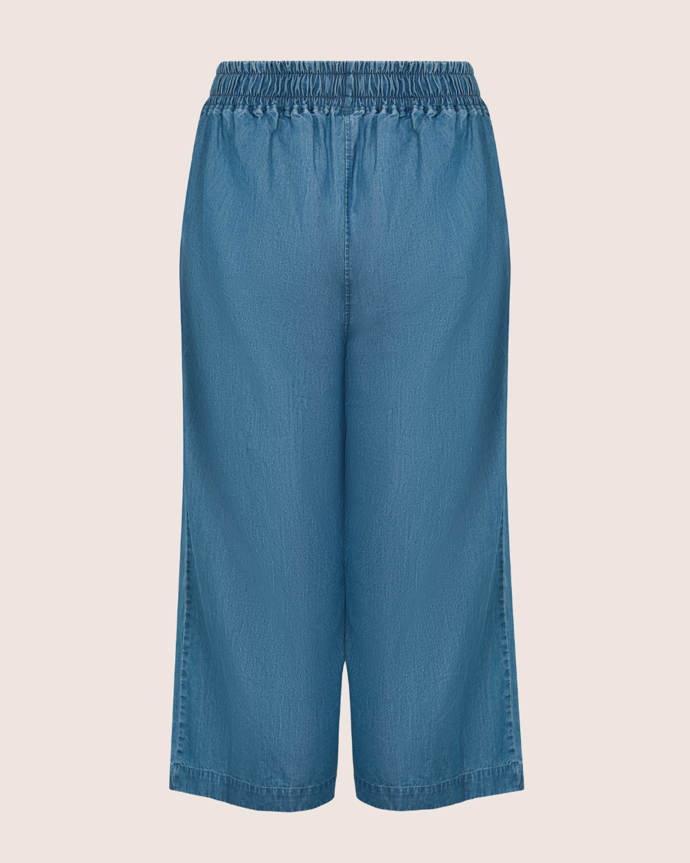 The Judy Washed Denim Blue Trousers feature a back view of wide-legged culottes crafted from organic cotton with an elastic waistband, set against a plain light background, merging comfort and style effortlessly.