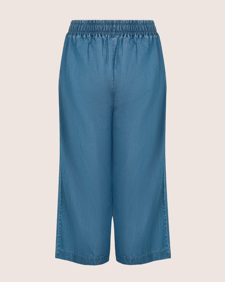 The Judy Washed Denim Blue Trousers feature a back view of wide-legged culottes crafted from organic cotton with an elastic waistband, set against a plain light background, merging comfort and style effortlessly.