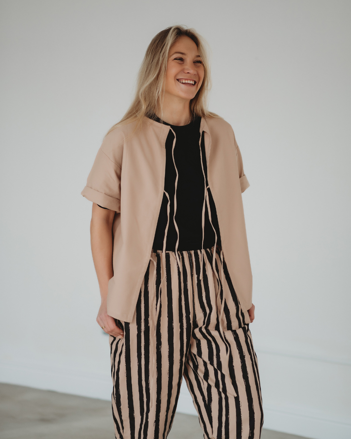 A person with long blonde hair smiles while wearing a beige jacket over a Doris Tie Back Clay Top, crafted from organic cotton. They stylishly pair it with beige and black striped pants against a plain, light-colored wall, highlighting versatile styling.