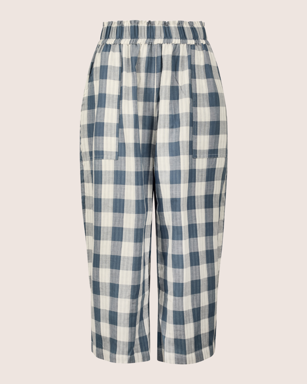 The Judy Reversible Blue Check Trousers are loose-fitting, high-waisted, featuring a blue and white checkered pattern with an elastic waistband, deep pockets, and wide legs for a relaxed style. The casual design is perfect against a neutral light beige background.