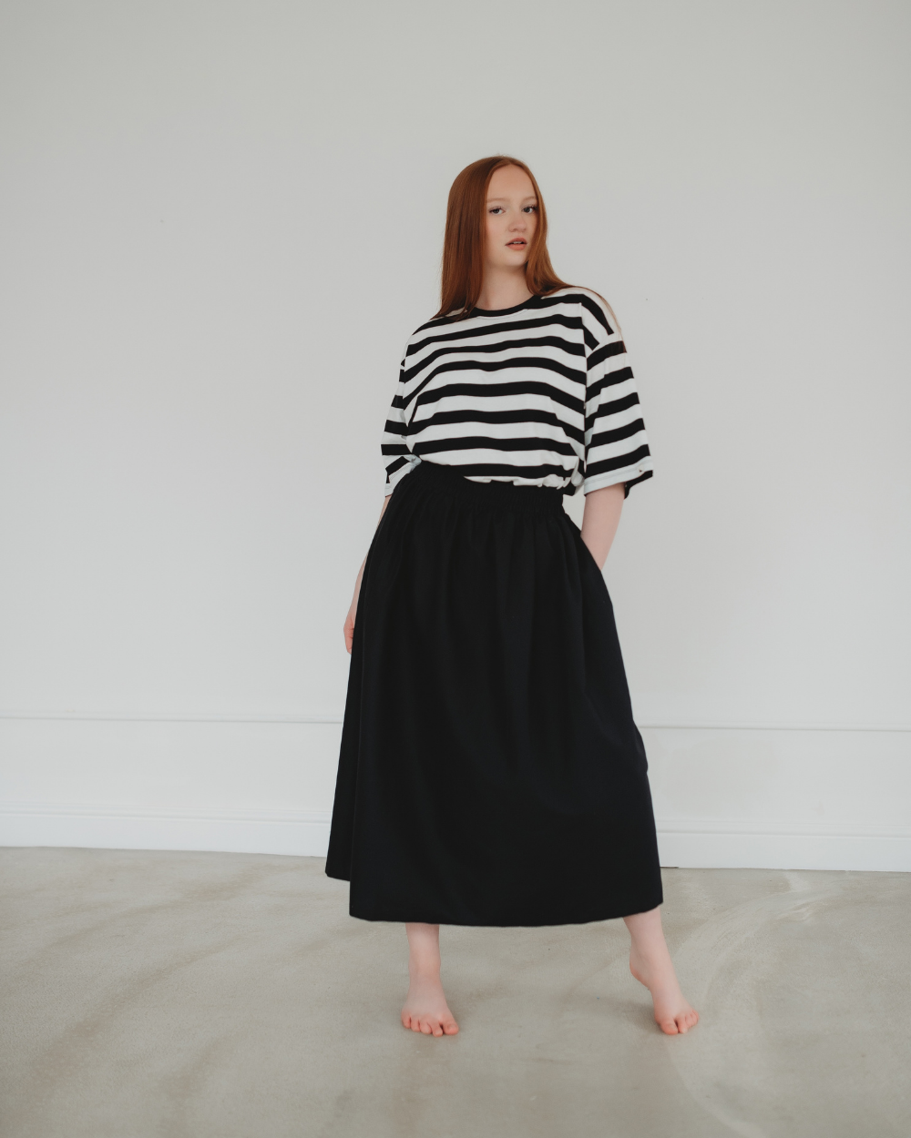 A person with long red hair stands barefoot on a light-colored floor, wearing an elasticated waistband black and white striped shirt paired with the versatile Joyce Easy Fit Black Skirt. They pose gracefully against a plain white wall, embodying the essence of a sustainable wardrobe.