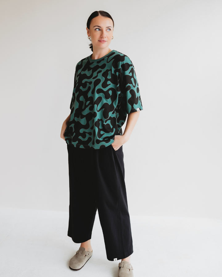 Against a white background, a person dons the Elsie Misty Print T-Shirt in vibrant green and black abstract patterns, paired with black wide-leg pants and beige shoes. Their short hair completes their look as they gaze to the side with hands in pockets.