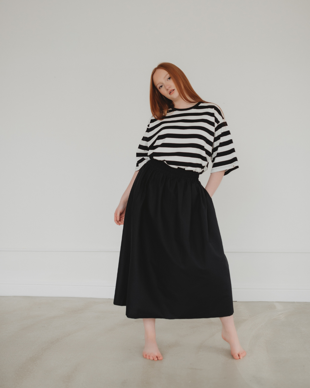 In a minimalist room, a person with long red hair stands barefoot, wearing a black and white striped shirt paired with the Joyce Easy Fit Black Skirt featuring an elasticated waistband. They pose gracefully against a plain white wall, effortlessly embodying sustainable wardrobe style.