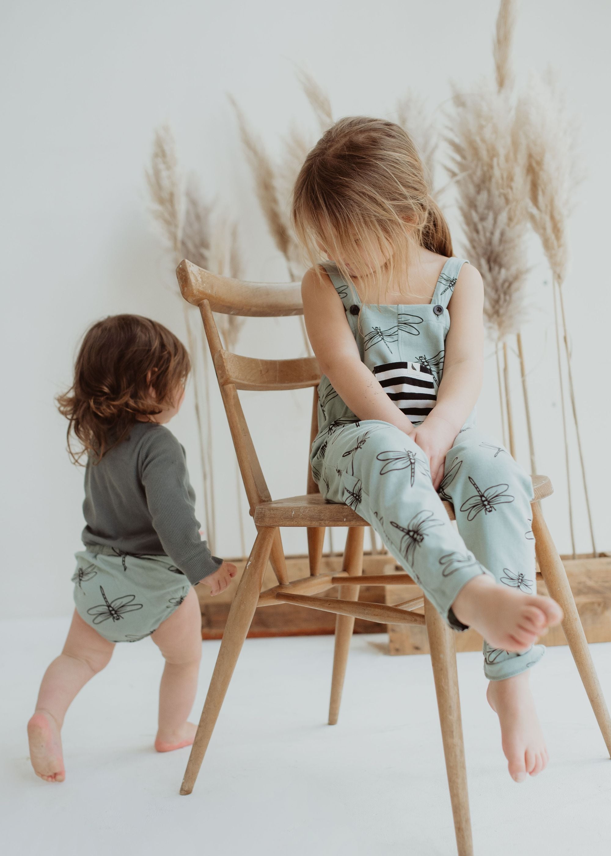 Child sits on a chair wearing turtledove dragonfly print organic cotton dungarees with stripe pocket. Other child wears the bloomer set of the same print. 