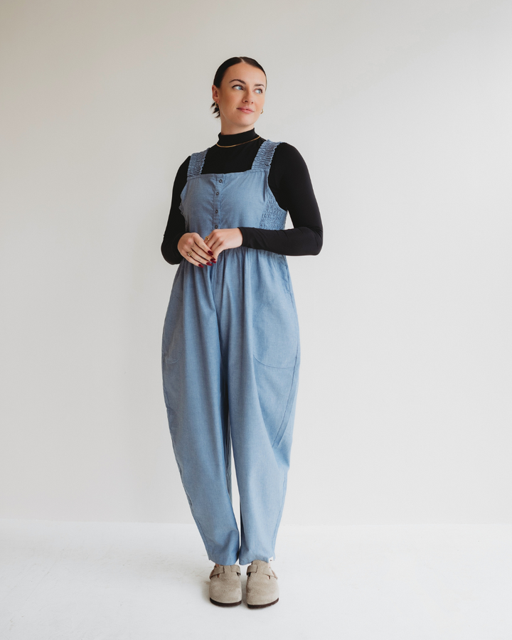 In a plain white setting, someone wears a black long-sleeve shirt beneath the organic Alice Chambray Dungarees paired with beige shoes. They have a thoughtful look, with hands clasped in front.