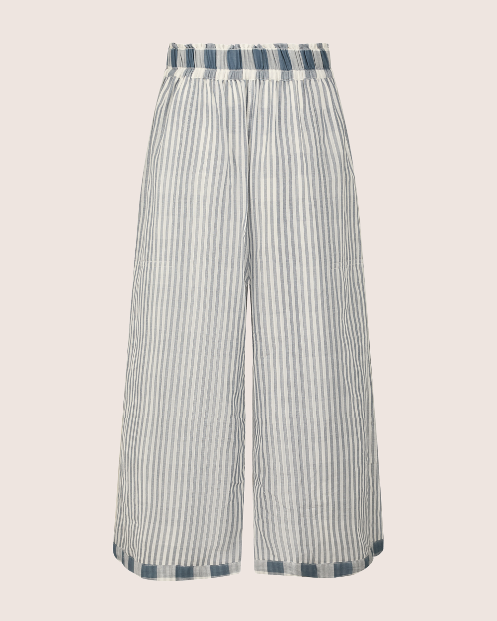 The Judy Reversible Blue Check Trousers are wide-legged, made from organic cotton, and feature a high elasticated waistband. They come in a lightweight fabric with vertical blue and white stripes, offering a relaxed fit and cropped length for effortless style.