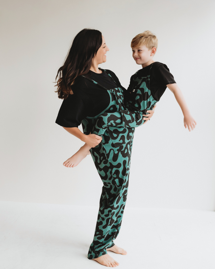 A woman and a young boy share smiles in matching Misty Print Dungarees made from organic cotton jersey. The woman holds him on a white floor against a plain backdrop, embracing their sustainable wardrobe.