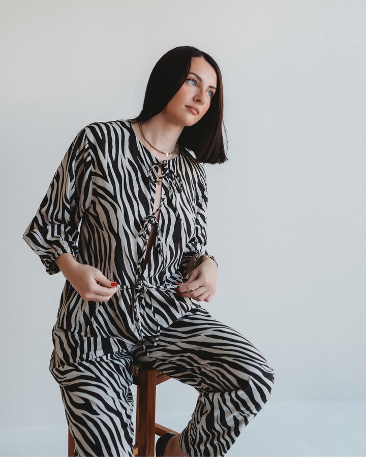 Seated on a stool, a woman with long black hair dons the eye-catching Blake Zebra Print Tie Blouse Top made from eco viscose. She gazes thoughtfully to the side against a plain white background.