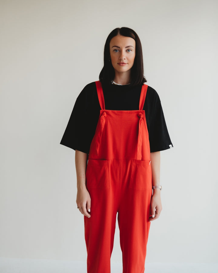 A person with long dark hair stands against a plain white background, wearing a black t-shirt and Lila Plain Red Dungarees in bright red organic cotton. They have a neutral expression and face the camera directly.