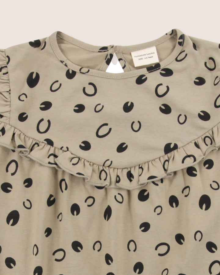 The Clip Clop Dress is a beige childrens dress featuring a Clip Clop print, round neckline, and ruffled trim across the chest. Made from 100% GOTS organic cotton, it has an easy fit with a small button at the back and visible clothing tags inside the collar.