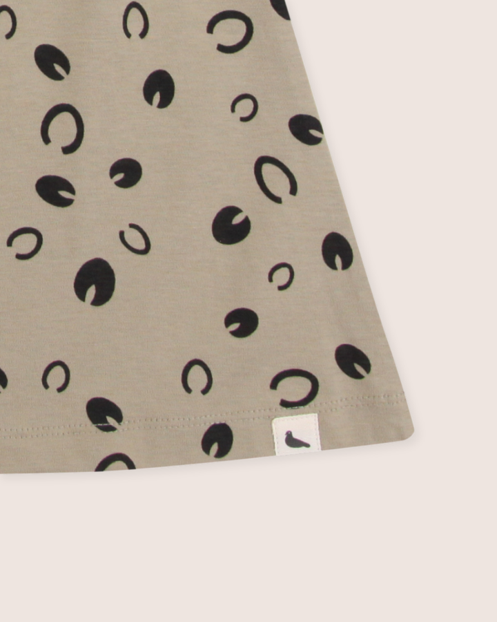 A close-up of beige fabric featuring scattered black circles and crescents, made from 100% GOTS organic cotton. A small white label with a black bird silhouette is sewn onto the bottom corner, akin to the Clip Clop Dress.