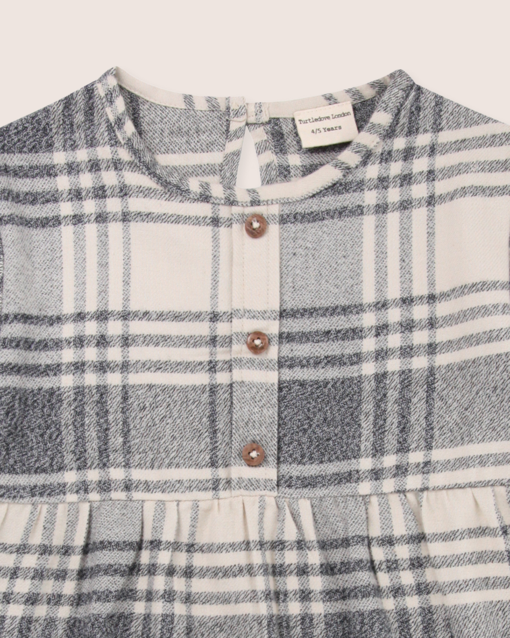 Close-up of the Brushed Check Dress in black, white, and gray plaid. Ideal for capsule wardrobes, it has a round neckline, front button-down with three brown buttons, and a gathered waist. Made from organic cotton with a back neckline tag.