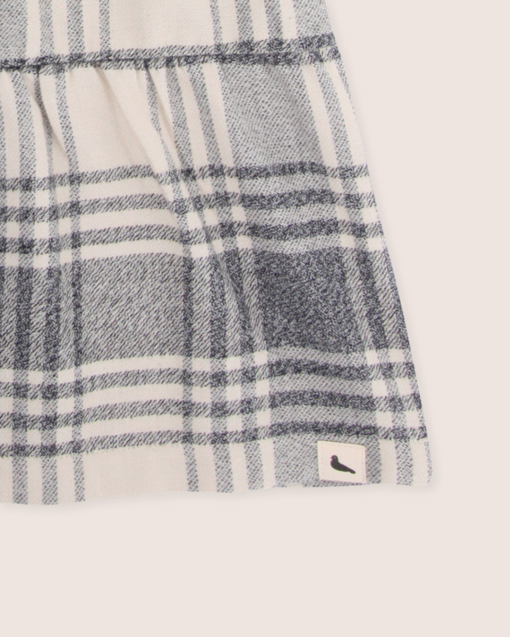 A textile close-up shows a black and white plaid perfect for capsule wardrobes. It has a mid-gathered section and a bottom-right label with a black bird silhouette, echoing the refined elegance of the organic cotton Brushed Check Dress.