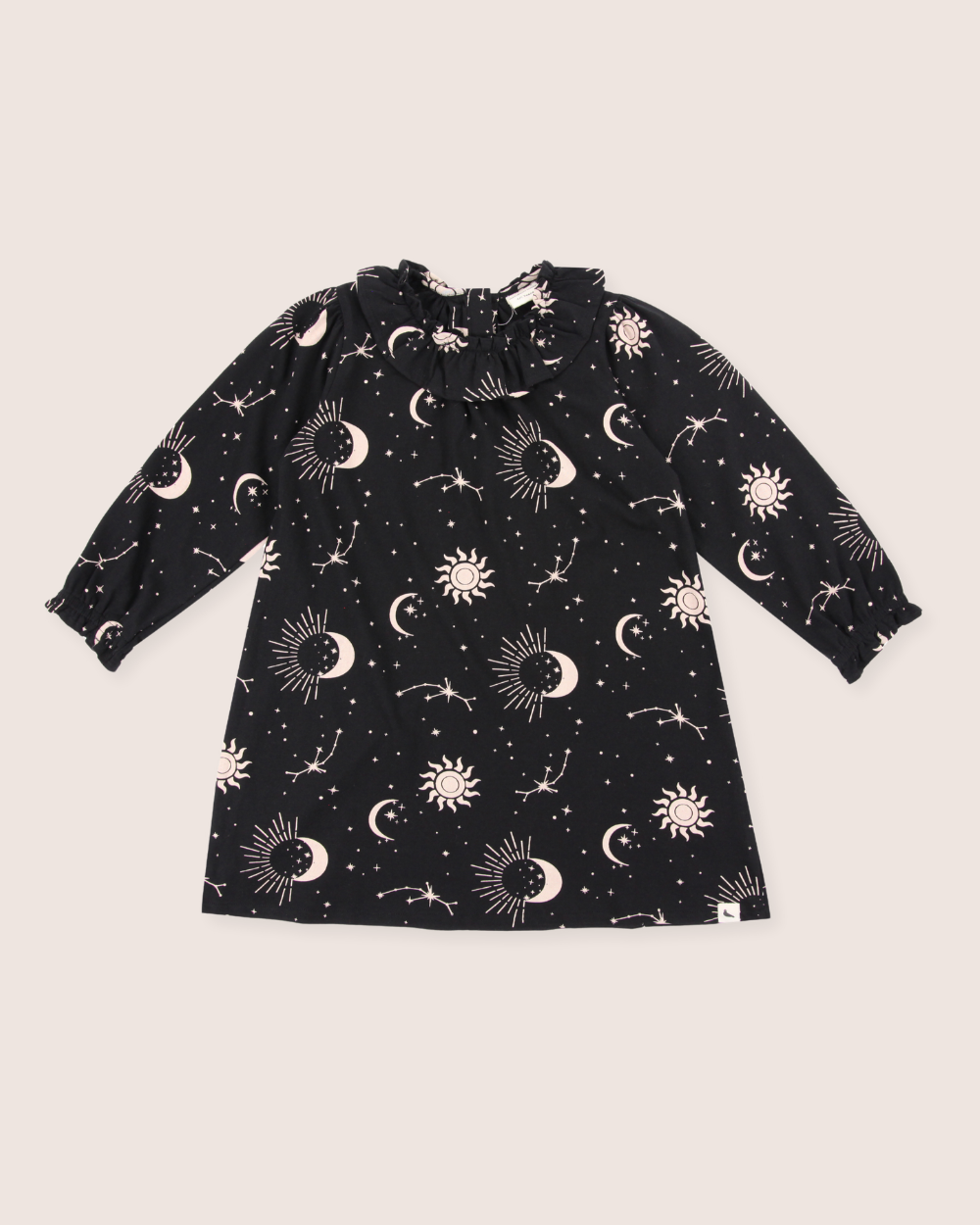 The Astrology Dress enchants with an astrology print on black organic cord fabric, featuring white sun, moon, and star motifs. It has long sleeves, a ruffled neckline, and a soft pinkish hue backdrop that perfectly blends style and whimsy for kids.