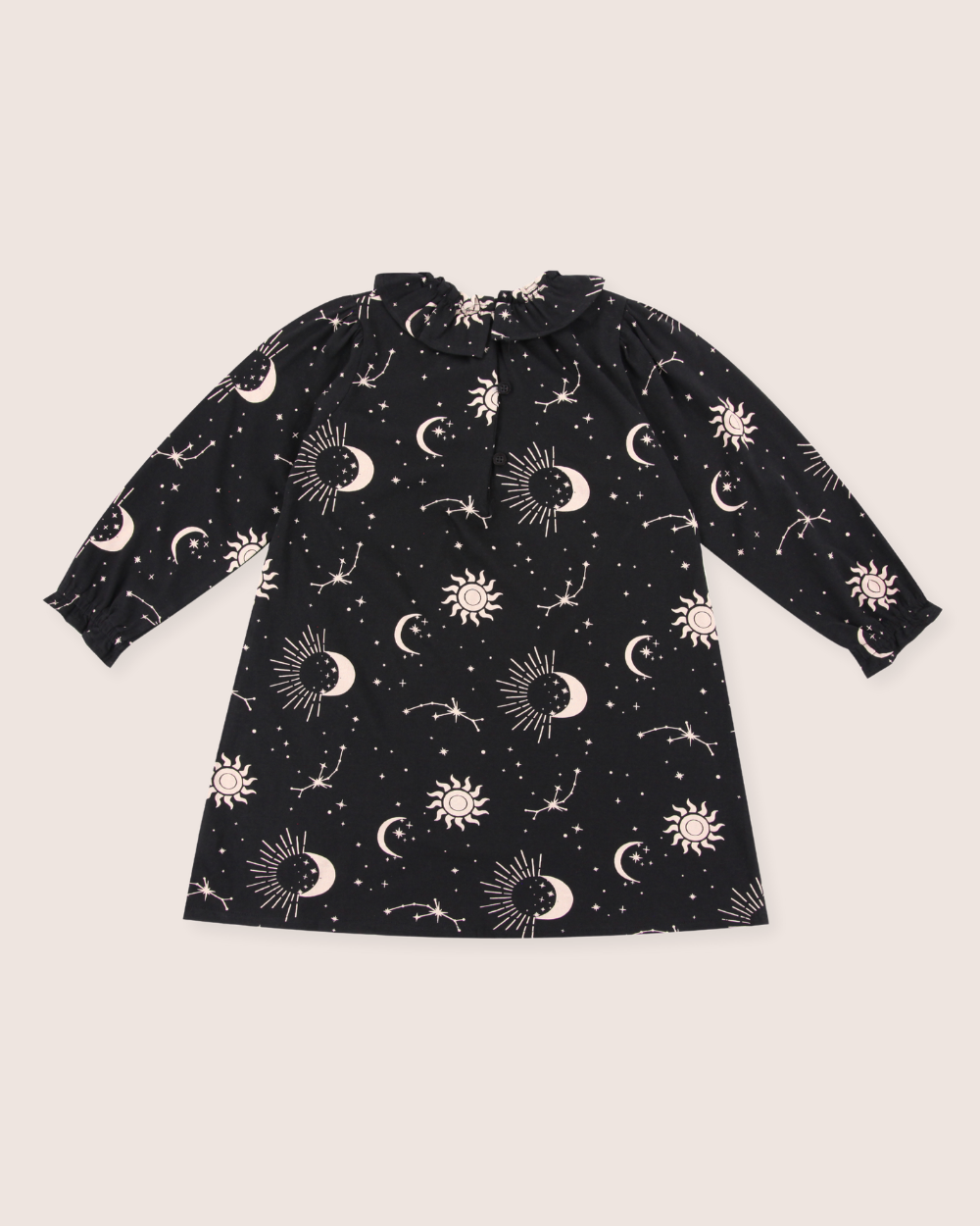 The Astrology Dress is a delightful kids garment made from organic cord fabric. It features a black long sleeve design with whimsical white celestial patterns of stars, moons, suns, and constellations on pale pink. The dress is styled with a gathered neckline and playful bow tie detail.