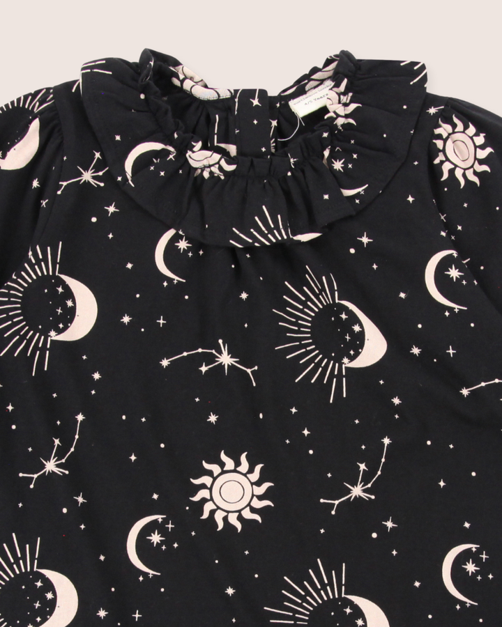 The Astrology Dress features black organic fabric with white crescent moons, stars, suns, and constellations print. It includes a celestial pattern and ruffled neckline, perfect for kids dresses.