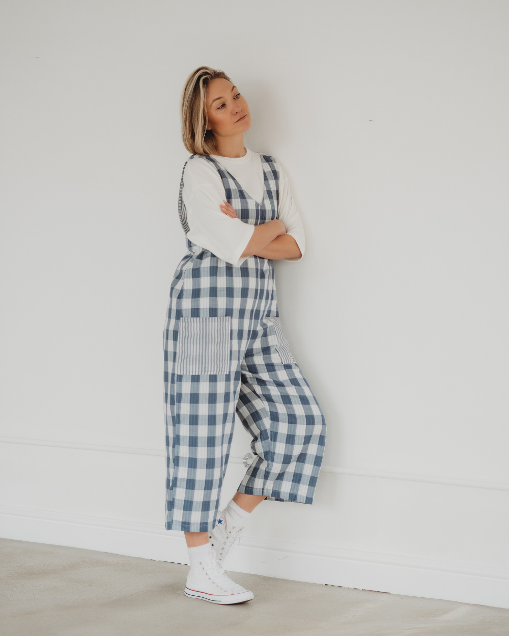 Leaning against a white wall with short blonde hair and white sneakers, the person exudes a relaxed vibe in their Grace Reversible Blue Check Dungarees, showcasing sustainable style with organic cotton.