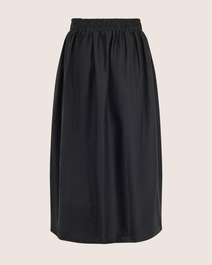 The Joyce Easy Fit Black Skirt, knee-length with an elasticated waistband, is displayed on a light beige background. Its relaxed fit and slightly gathered design make it ideal for a sustainable wardrobe, offering both casual comfort and style.