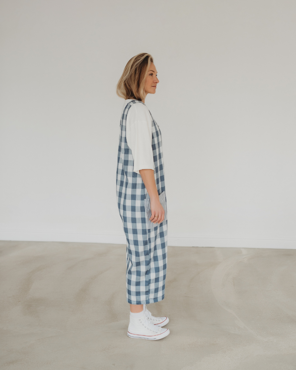 A person stands in profile against a plain background, wearing Grace Reversible Blue Check Dungarees crafted from organic cotton over a white shirt and white sneakers. The checkered jumpsuit exudes a casual, relaxed style, perfect for a sustainable wardrobe.